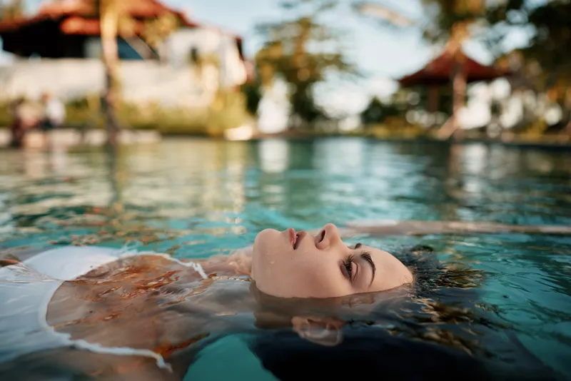A person floats in a clear pool with eyes closed, savoring relaxation and calm. Outdoor setting with sunlit water and resort vibe, a moment of wellness and rest.