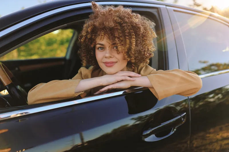 Smiling woman with curly hair leaning on a car window, casual lifestyle scene, warm daylight, retro film color, contemporary portrait conveying confidence and carefree mood.