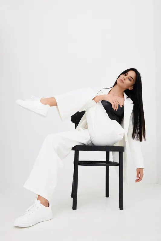 Stylish woman in white wide-leg pants and sneakers poses sitting on chair with relaxed expression against plain light background in modern minimalistic fashion portrait.