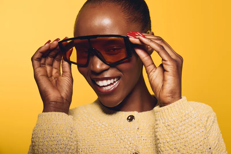 Happy African woman with dark skin wearing oversized sunglasses and yellow sweater smiles joyfully on yellow background. Concept of fashion, beauty, confidence, and vibrant style.