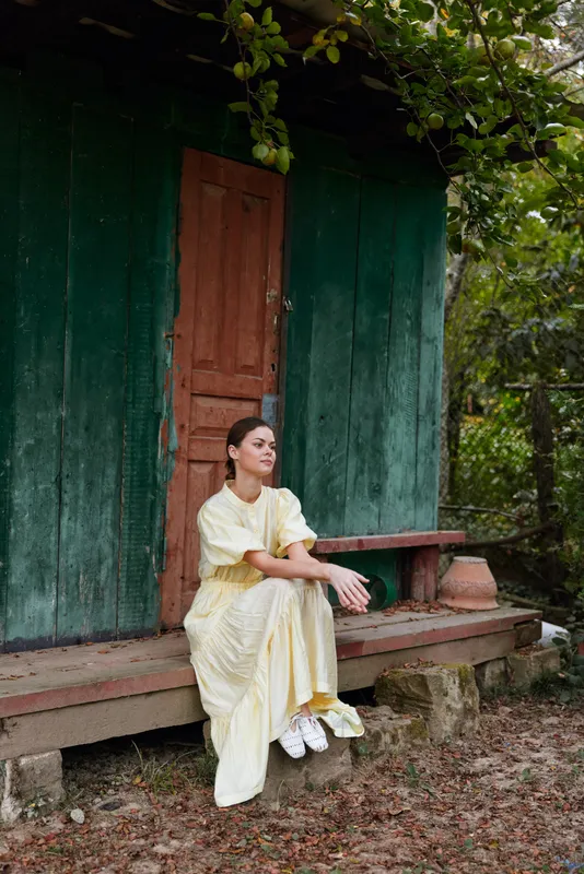 Woman in a flowing yellow dress sits on a bench beside a rustic green doorway, outdoors among trees, conveying calm, contemplative mood and a simple natural lifestyle.
