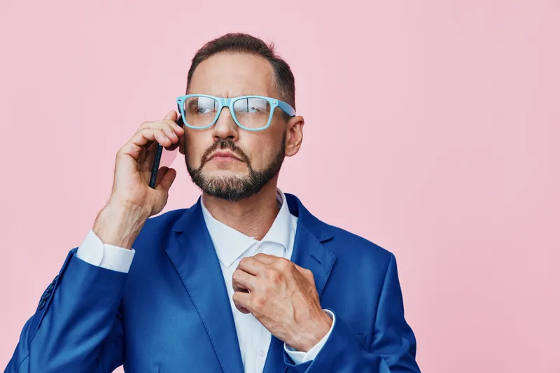Professional man in a blue suit speaks on a mobile phone against a solid pink background, conveying focus, confidence, and determination in a modern business scene.