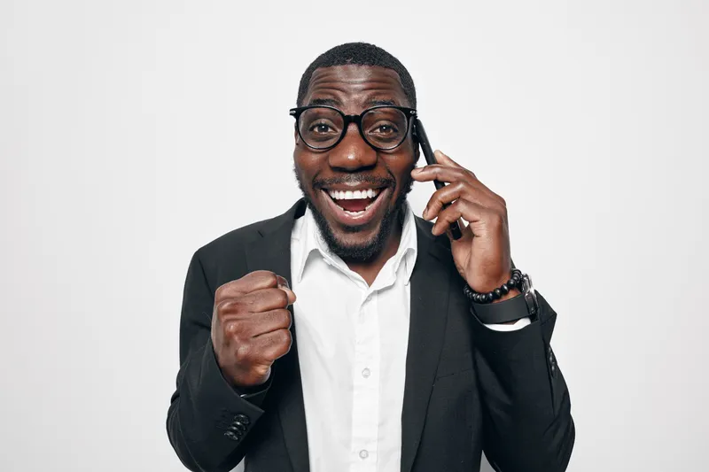 African American man with dark skin wearing glasses and suit talking on phone with happy expression and fist pump gesture on white background.