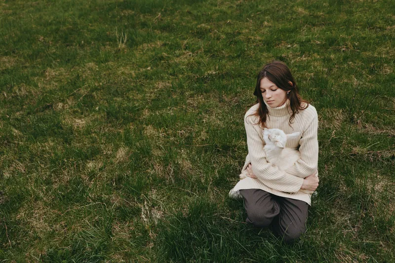 Lifestyle moment with a woman outdoors on grass, kneeling and holding a goat kid, cozy sweater, calm smile, natural light, soft breeze, authentic connection, warm colors, tranquil