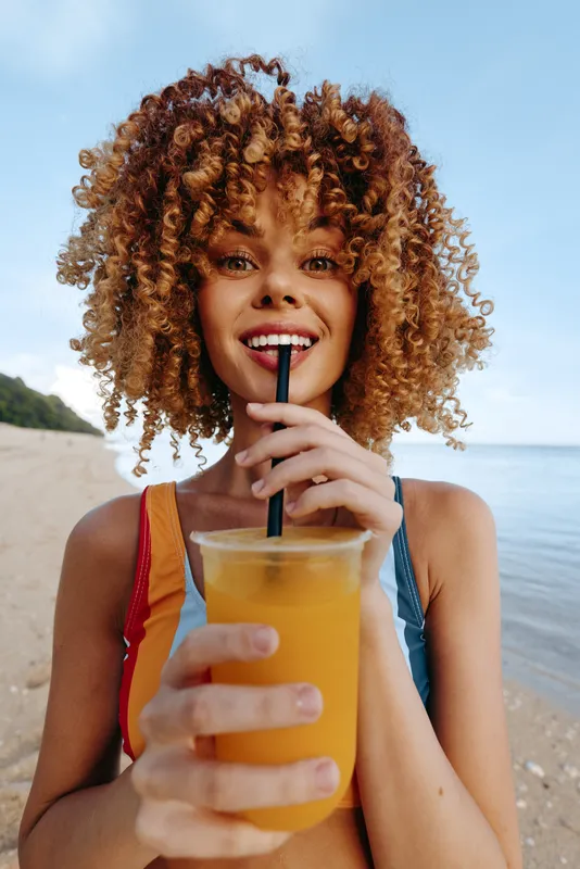 A smiling young woman with curly hair drinks orange juice through a straw on a sunny beach, wearing a two piece swimsuit, capturing a carefree summer moment.