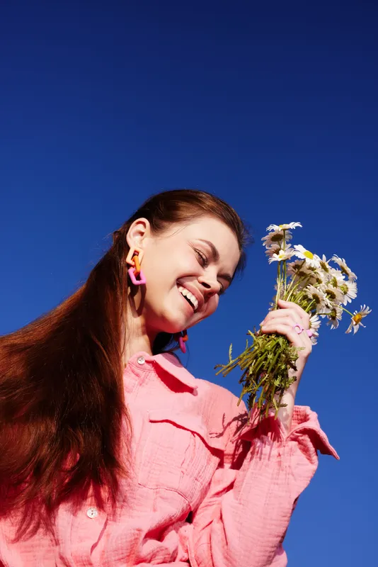 Young woman smiling brightly while holding a bouquet of daisies against a clear blue sky, dressed in a pink shirt, exuding joy and a sense of spring