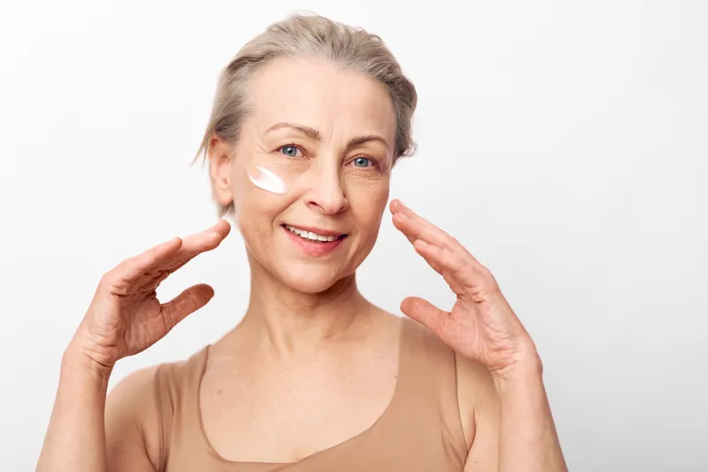 Mature woman with light skin applies facial cream with hands near face, smiling gently. Concept of skincare, aging beauty, self-care, and natural wellness on plain background.