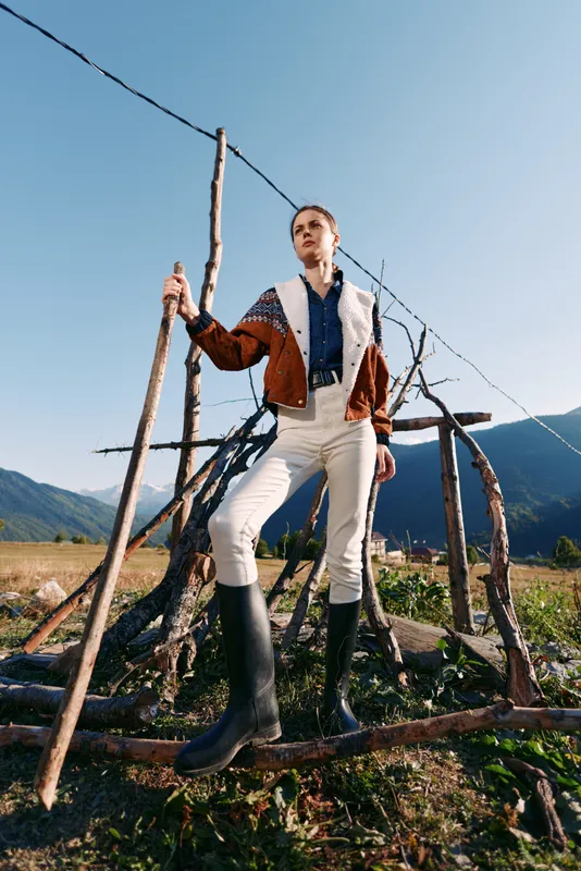 Woman in rubber boots holds a wooden stick standing by a rustic fence in countryside, wearing patterned jacket and white trousers, posing confidently against mountains and clear sky.