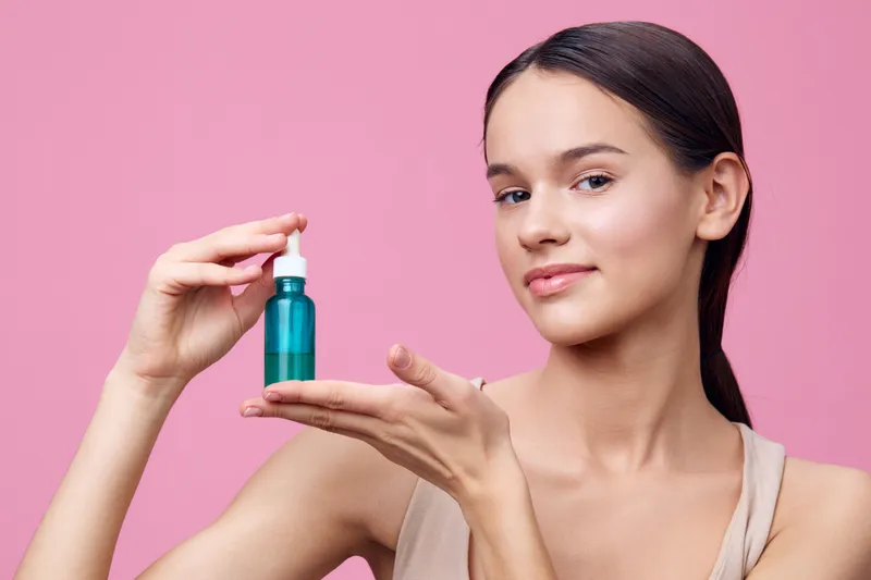 Young woman showcasing a skincare serum in a teal bottle against a soft pink background, embodying beauty, skincare, and self care, with a serene expression