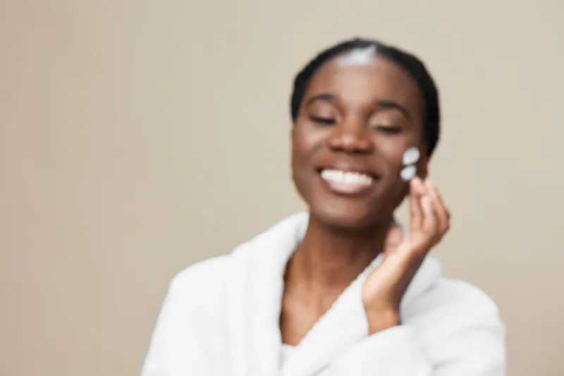 Smiling Black woman applying skincare cream in a cozy spa setting, promoting self care and wellness with a neutral background and soft lighting