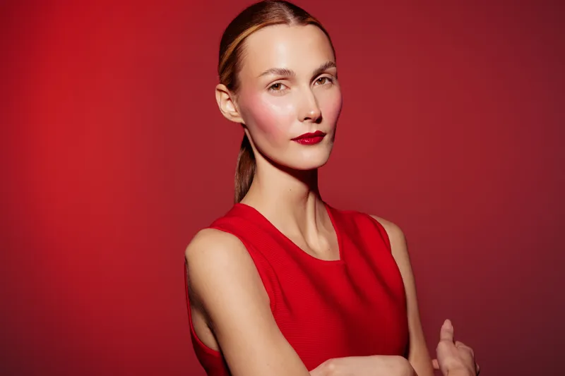 Confident woman in red sleeveless dress poses with arms crossed against red background. Studio portrait with natural makeup and sleek hairstyle. Concept of elegance, fashion, and confidence.