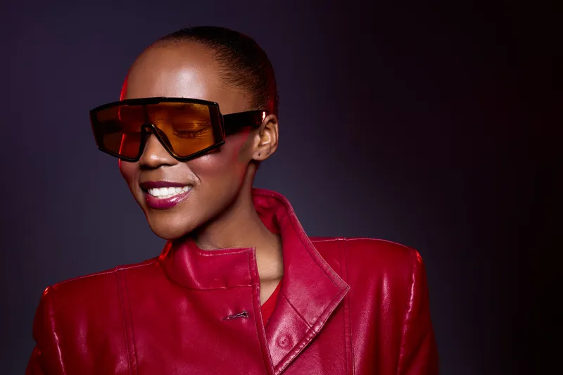 Smiling African American woman with dark skin wearing oversized brown sunglasses and a red leather jacket. Studio portrait with dark background. Concept of fashion, confidence, and style.