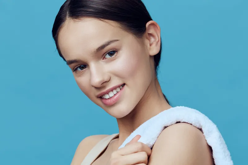 Smiling young woman with a towel on her shoulder against a blue background, showcasing healthy skin and a fresh, vibrant look, ideal for wellness and beauty concepts