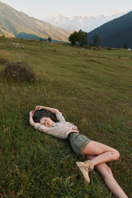 Young woman rests on a grassy hillside in a scenic valley, eyes closed and arms stretched overhead, enjoying calm after a hike in bright daylight.