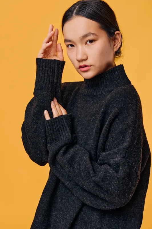 Young woman with dark hair wearing a black sweater poses thoughtfully against orange background. Studio portrait of model with natural makeup and casual cozy fashion style.