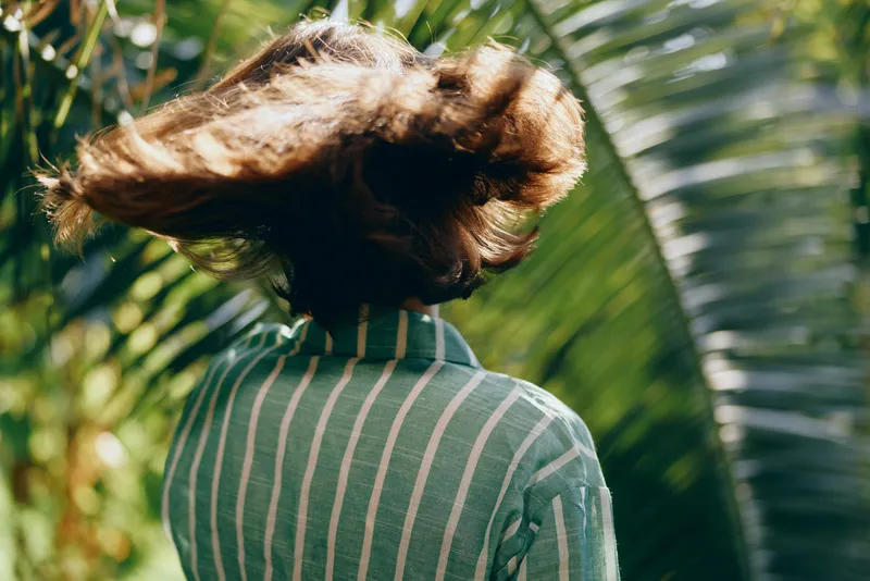 Woman outdoors, back view, striped shirt, wind blows hair, lush greenery, sunny day, fashion moment, natural light, casual elegance, movement, fresh energy, relaxed mood, candid style, outdoor