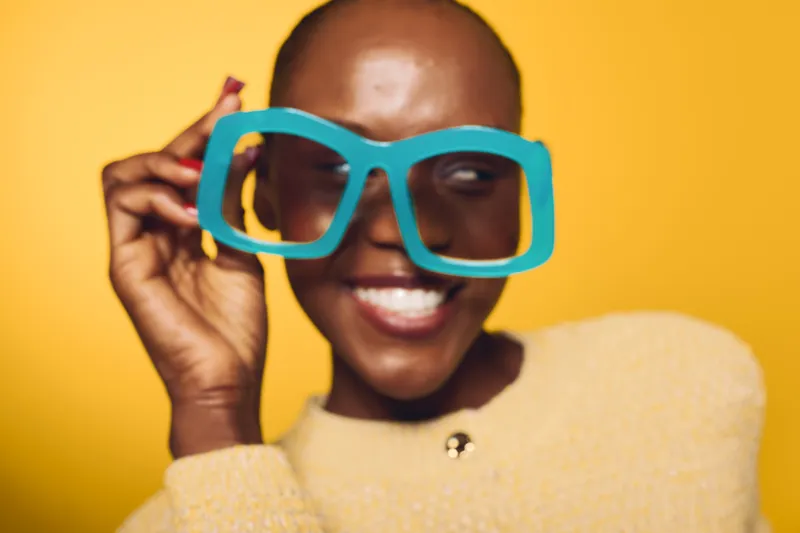 Smiling happy black woman with dark skin wearing large blue glasses and yellow sweater. Close-up portrait on yellow background. Concept of fashion, joy, and modern style.