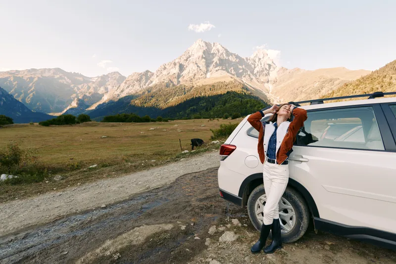 Woman leans against white car by a dirt road in mountains, nature landscape for travel and adventure. Relaxed portrait with jacket and boots enjoying scenic meadow and sunlight.