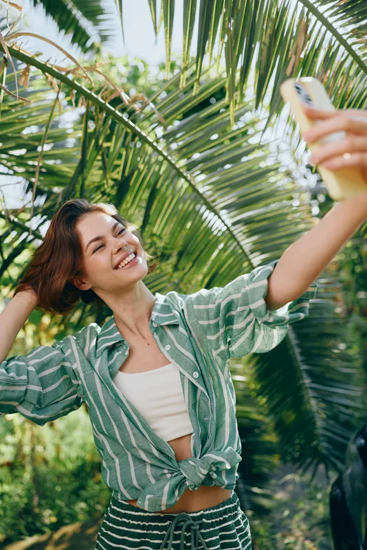 Bright portrait of a smiling woman taking a selfie among tropical palms, wearing a green striped shirt and white top.