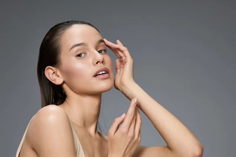 Elegant young woman with smooth skin applying skincare products, showcasing a natural beauty concept against a soft gray background