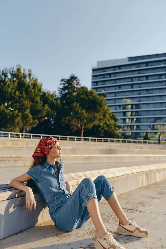 Modern boho-western denim street style woman relaxing outdoors in casual outfit with red headscarf, enjoying sunny day with film color warm tones and natural background.