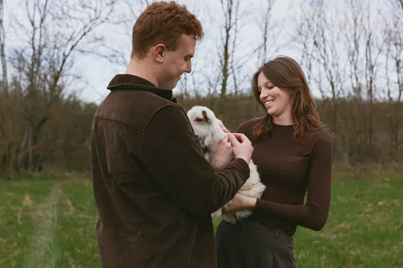 Couple outdoors with goat kid, sharing a gentle moment in a grassy field, capturing a warm lifestyle scene with a candid, natural feel and genuine connection between people and animal companions.