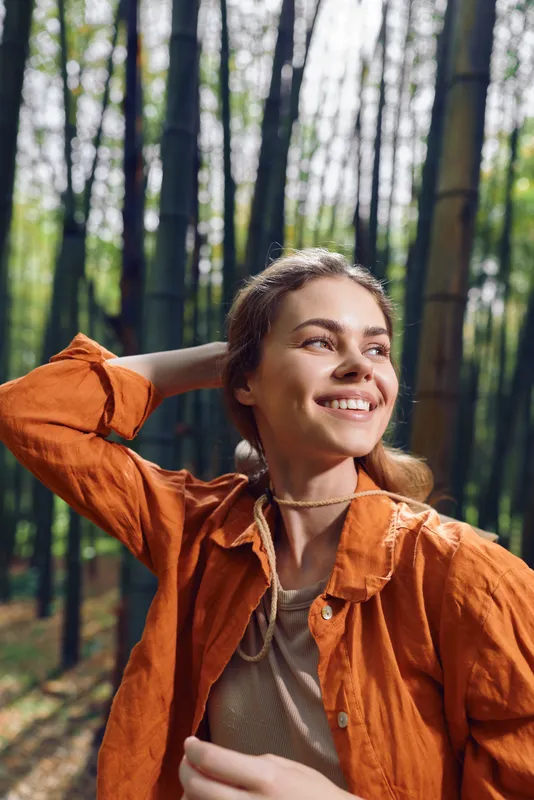 Woman smile portrait in bamboo forest, orange jacket and happy nature vibe, young female looking up with hand behind head, sunlight filtering through tall trees in outdoor scene