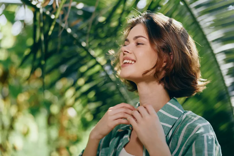 A young woman with short brown hair smiles softly in a sunlit tropical garden, wearing a striped green shirt.