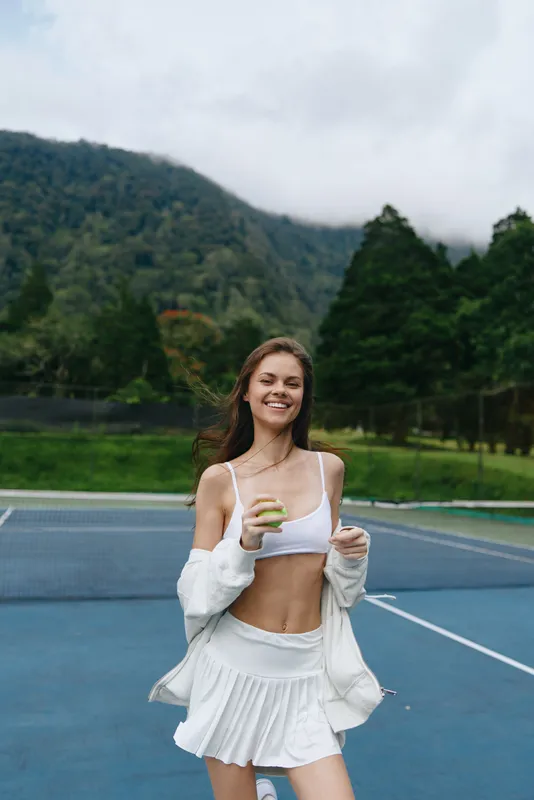 Joyful young woman in sporty attire enjoying tennis on an outdoor court with mountains in the background, embodying a healthy lifestyle and athletic spirit