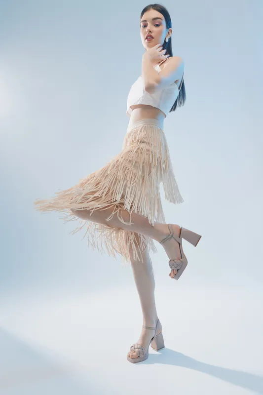 Young woman in a beige fringed skirt and white crop top poses elegantly on one leg against a light background. Concept of woman beauty fashion and style.