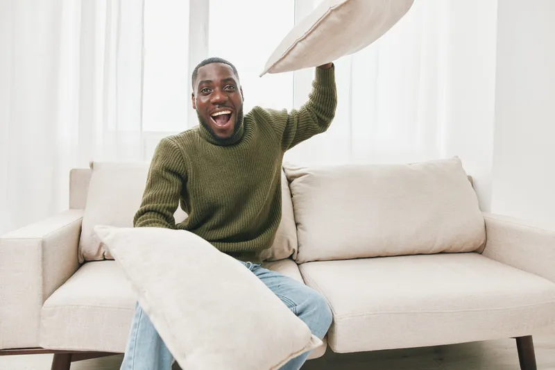 A joyful Black man playfully holding pillows, dressed in a cozy green sweater, sitting on a light colored couch in a bright, minimalistic room, promoting fun and relaxation