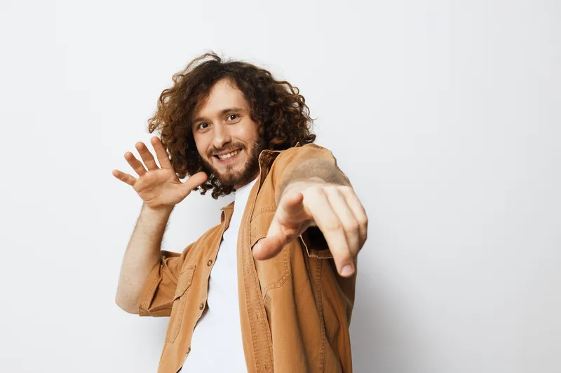 Cheerful young man with curly hair in casual clothing, posing playfully against a white background, expressing joy and energy, perfect for lifestyle concepts