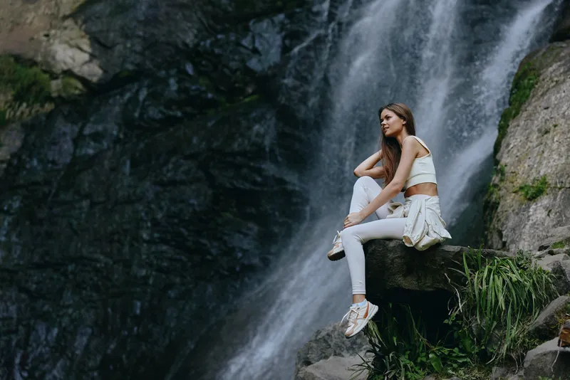 Young woman relaxing near a waterfall, wearing white casual clothes and sneakers, enjoying nature in a peaceful outdoor environment with rocky background and flowing water.