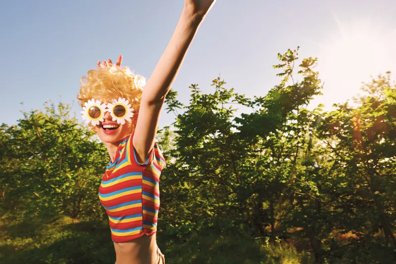 A joyful woman in a striped top raises her arm in the sun, wearing a playful sunflower mask. An outdoor scene with bright retro film lighting and vivid colors.