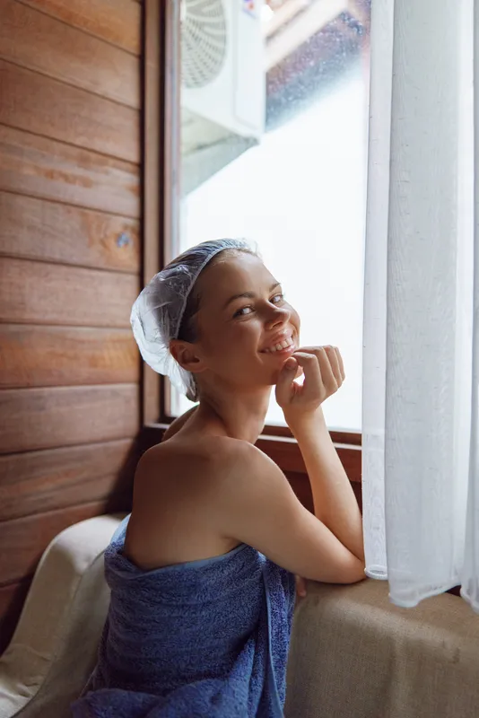 young woman wrapped in towel relaxing indoors near window with wooden wall background and natural light, enjoying calm and peaceful moment after shower
