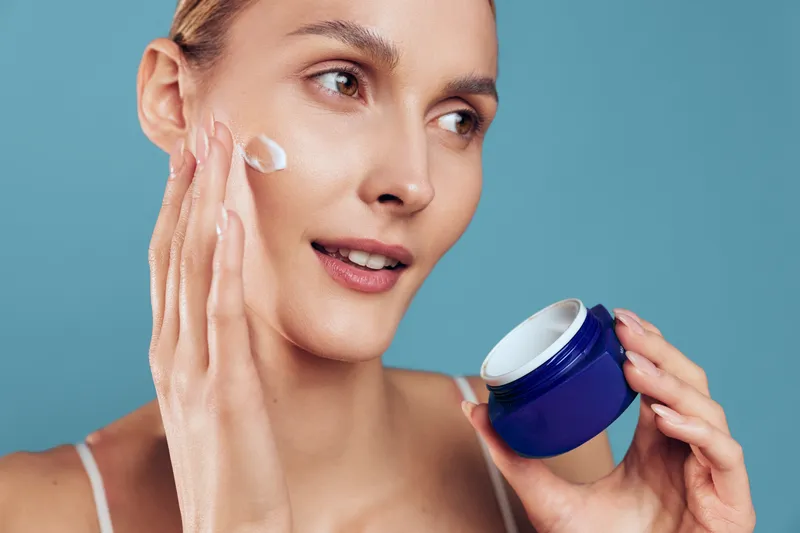 Young woman skincare beauty moment with cream on cheek, applying face moisturizer while holding a cosmetic jar, clean blue background, calm routine concept for hygiene and self care