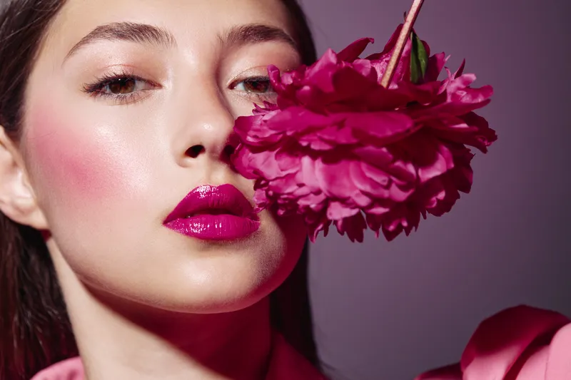 Close-up of a young woman with glossy bright pink lipstick and glowing skin holding a vibrant pink peony near her face. Soft lighting accentuates the beauty and fashion concept.