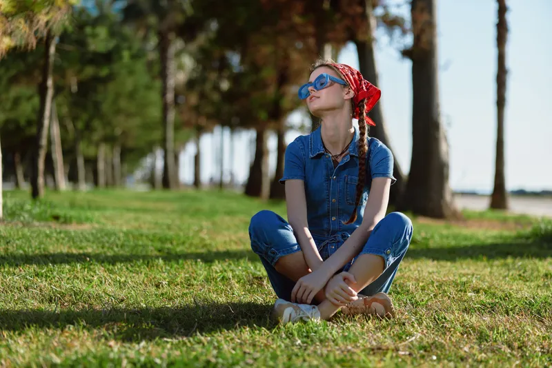 lifestyle woman in modern boho-western denim street style sitting on grass with red bandana and sunglasses in natural sunlight, film color effects enhance mood
