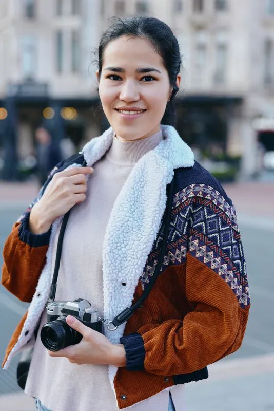 Camera in hand, female photographer on a city street, wearing a knit sweater and colorful jacket, capturing candid urban moments with confident, creative vision and approachable charm
