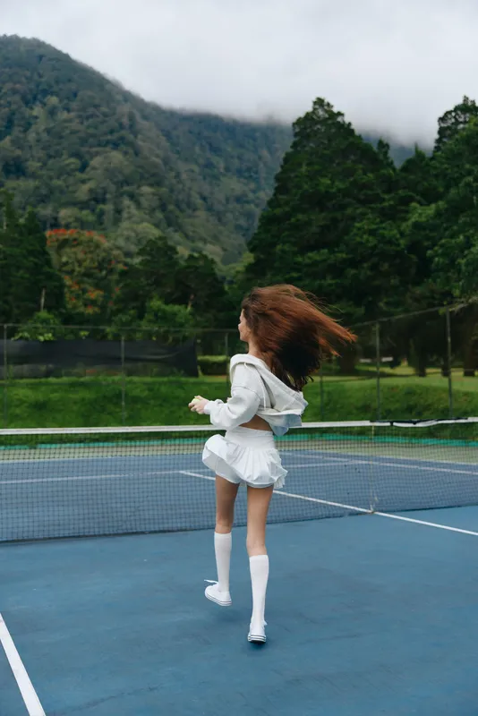 Young female athlete in white sportswear practicing tennis against a lush green mountain backdrop, embodying energy and focus in a vibrant, seasonal setting