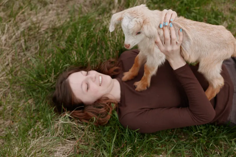 A woman lies on grassy ground outdoors, smiling while holding a young goat kid. The scene conveys casual, playful lifestyle and warm, gentle animal bonding in a sunny, natural setting.