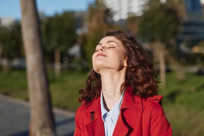 Young woman with curly hair enjoys fresh air outdoors wearing a red coat and white shirt with closed eyes and relaxed expression on a sunny day.