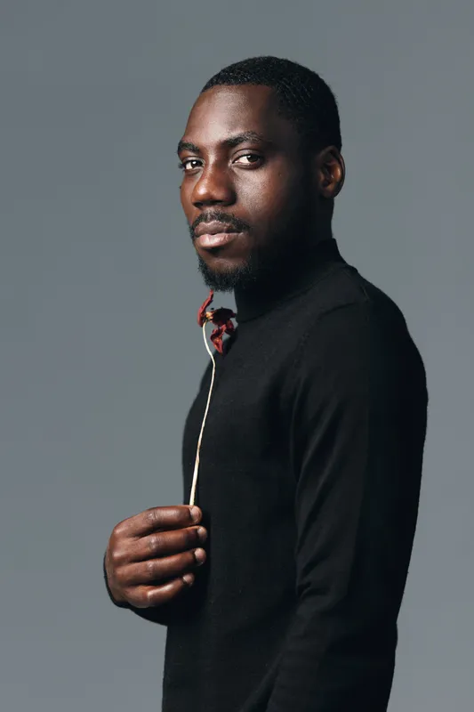 African man with beard in black sweater holding dried flower looking at camera in creative conceptual visual style studio portrait with neutral gray background and serious expression.