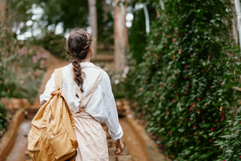 Young woman walking on a garden path with a backpack, wearing a light shirt and suspenders, surrounded by greenery and colorful flowers, evoking a sense of adventure and exploration