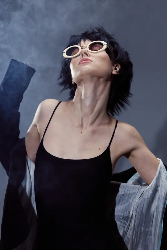 Stylish woman in sunglasses wearing black dress with open shirt on smoky background, posing confidently with modern fashion vibes.
