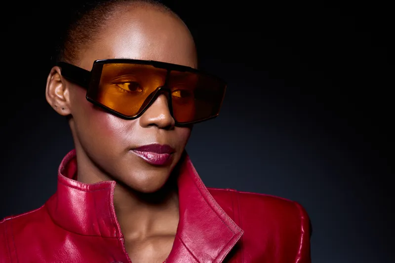 Portrait of confident black woman with dark skin wearing oversized amber sunglasses and red leather jacket. Studio shot with dramatic lighting highlights fashion and style concept.