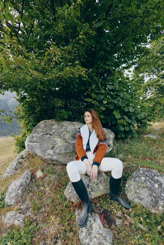 Woman sitting on mossy rocks under a large tree in nature, wearing boots and casual coat, outdoors portrait capturing relaxed style, rural landscape and serene autumn mood.