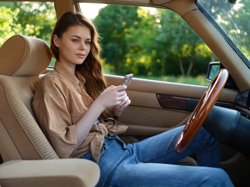 Young woman texting in a vintage car, wearing casual outfit, exuding relaxed vibes in a lush green setting, ideal for lifestyle and travel themes.