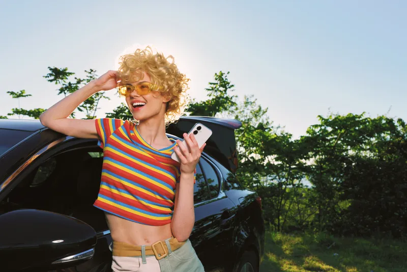 Woman with curly blonde hair in a striped crop top smiles beside a car, holding a phone, sunlit outdoor scene, exuding a cheerful retro lifestyle and carefree mood.