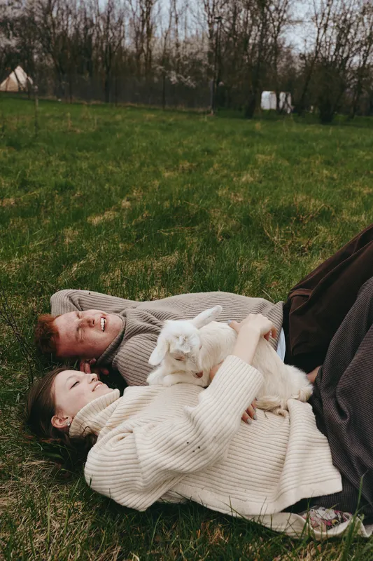 Charming couple outdoors, lying on grass, cozy sweaters, a playful goat kid resting between them, sunlit moment, relaxed mood, natural colors, rural countryside background